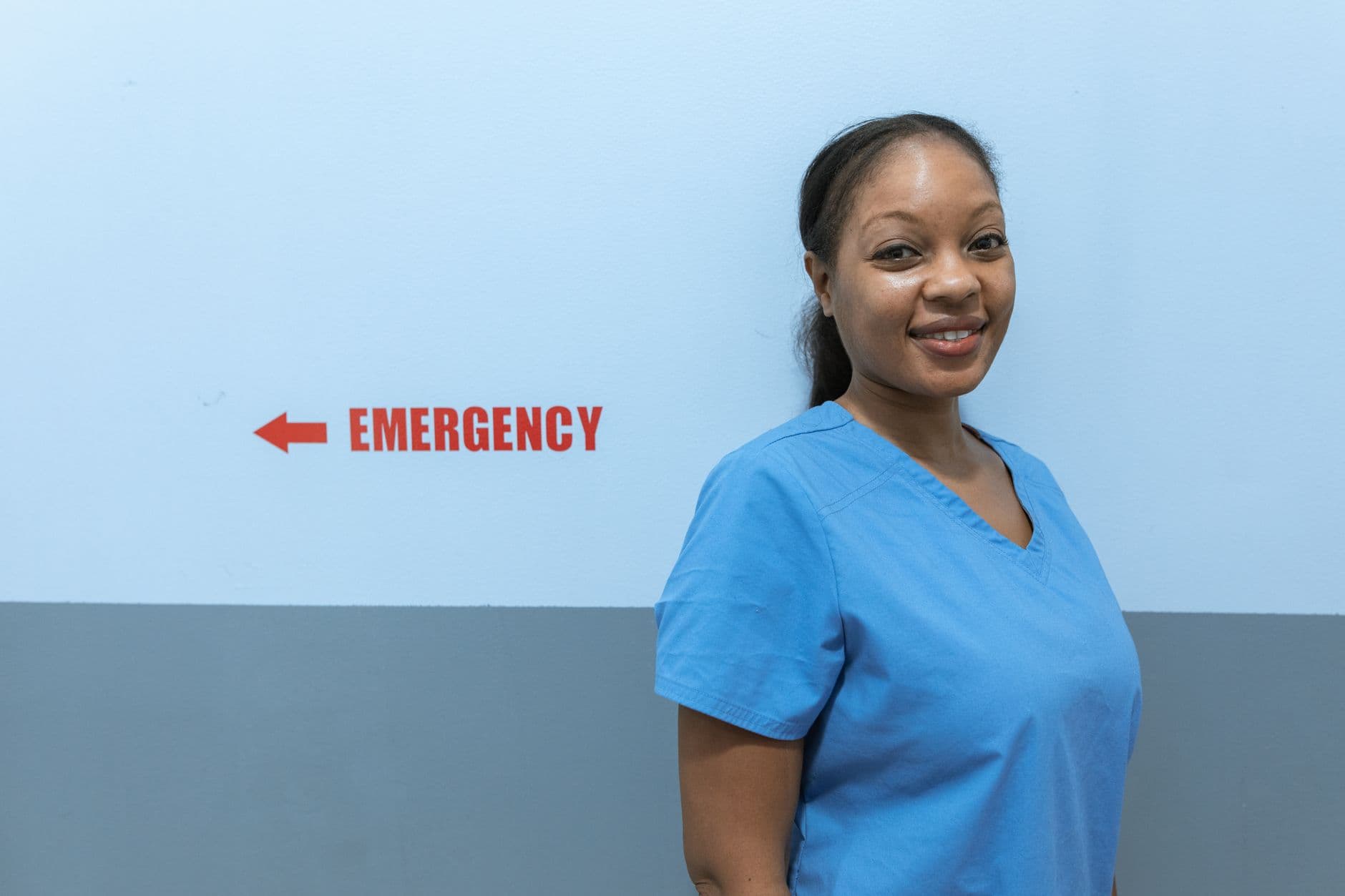 Happy healthcare worker in scrubs standing near emergency room sign.