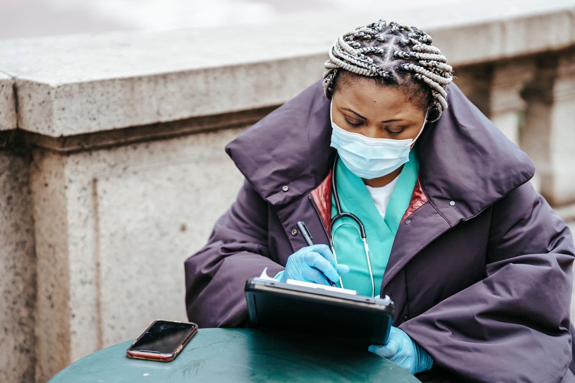 Middle aged African American female medic in outerwear and mask taking notes on paper at table with cellphone in city